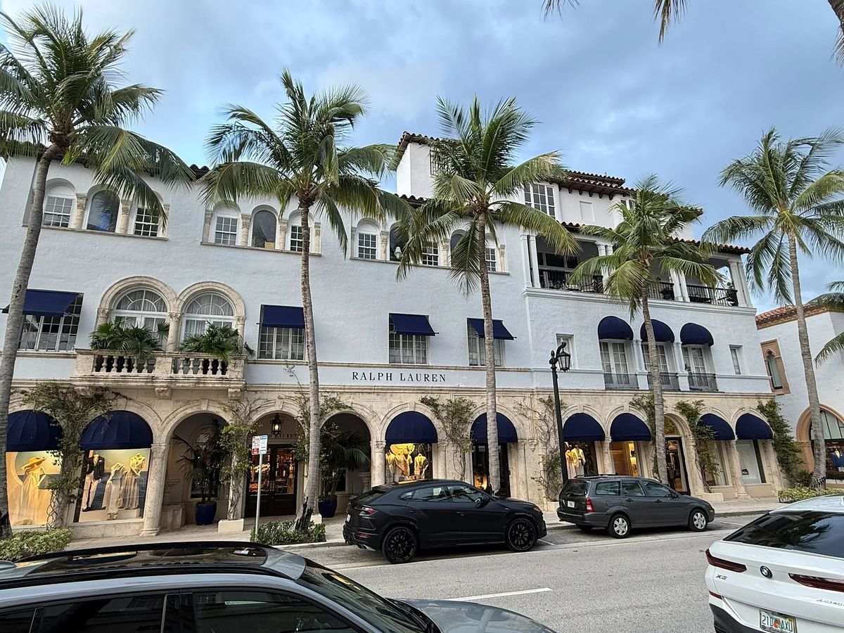 Building facade featuring arched windows, balconies, covered awnings, and palm trees.