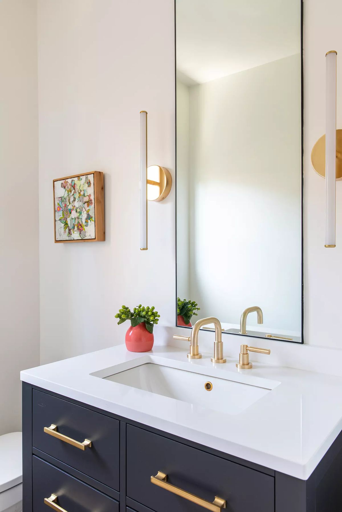 Bathroom with dark vanity, white countertop, mirror, artwork, potted plant, and gold wall sconces.
