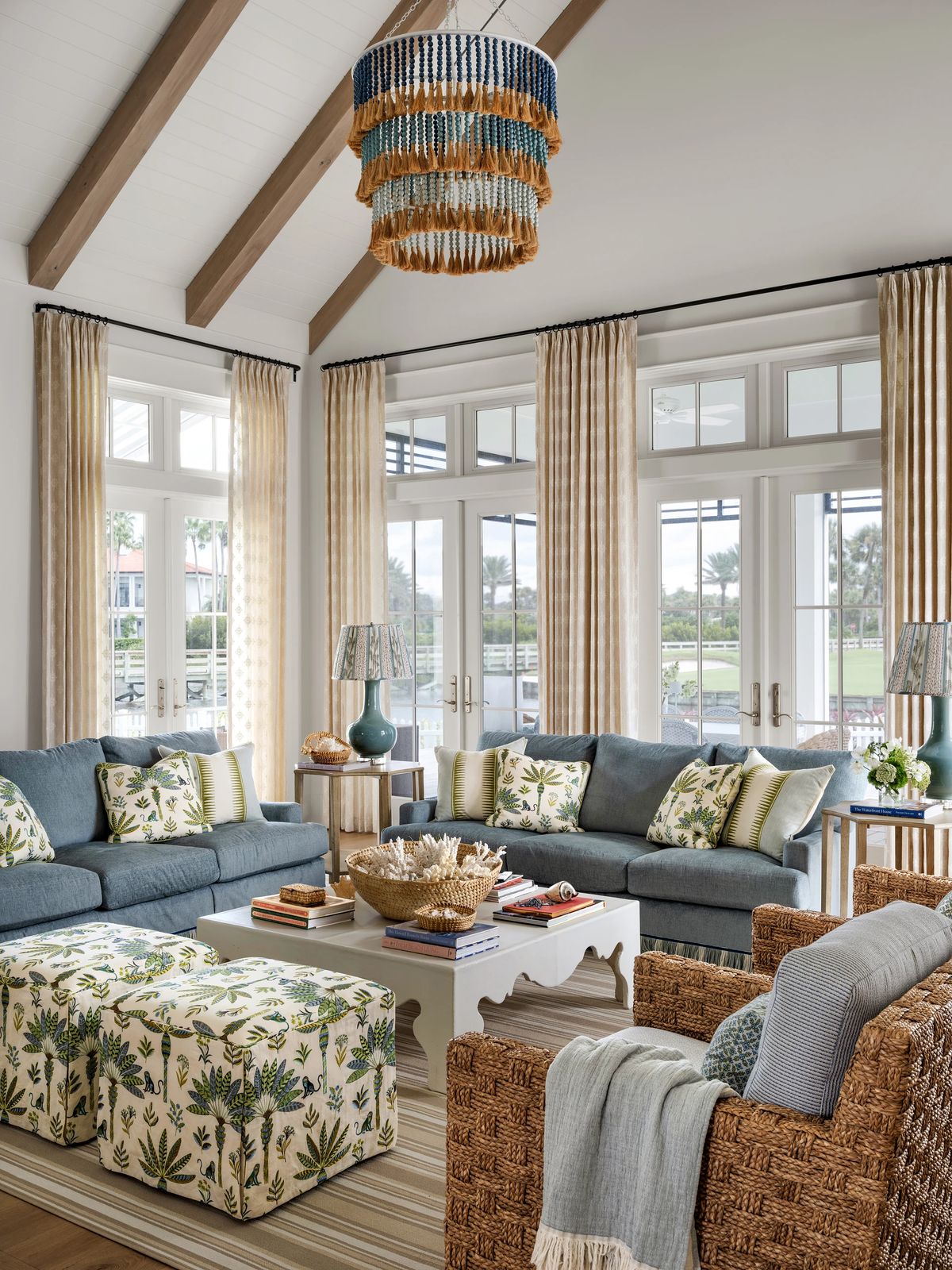 Living room with blue sofas, floral pillows, coffee table, wicker armchair, large windows, and chandelier.