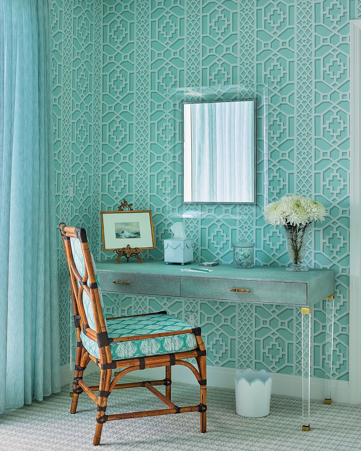 Office with turquoise patterned wallpaper, glass desk, wooden chair, mirror, flowers, and blue curtains.