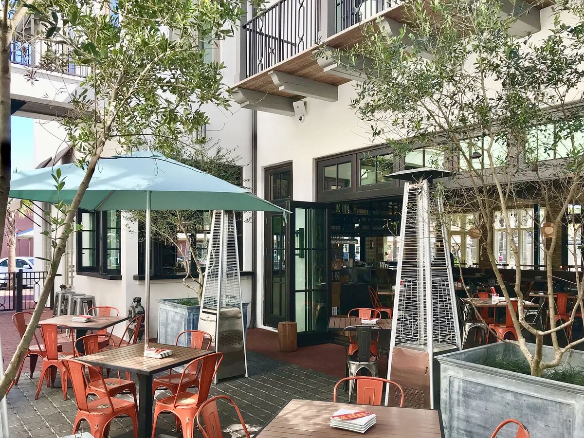 Outdoor patio with metal tables, chairs, turquoise umbrella, heat lamps, and potted trees.