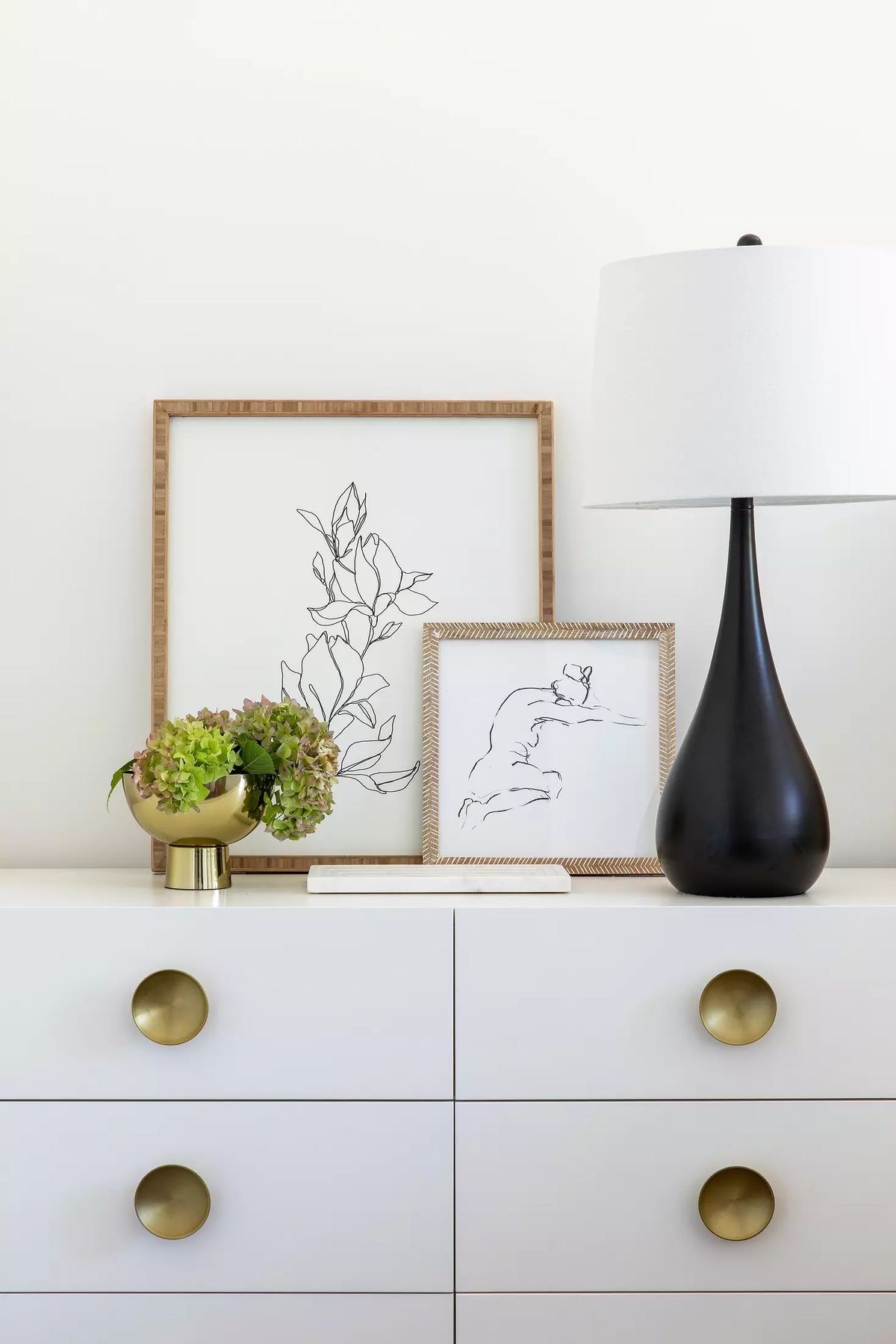 Tabletop with white dresser, black lamp, green plant, and framed artworks against a white wall.