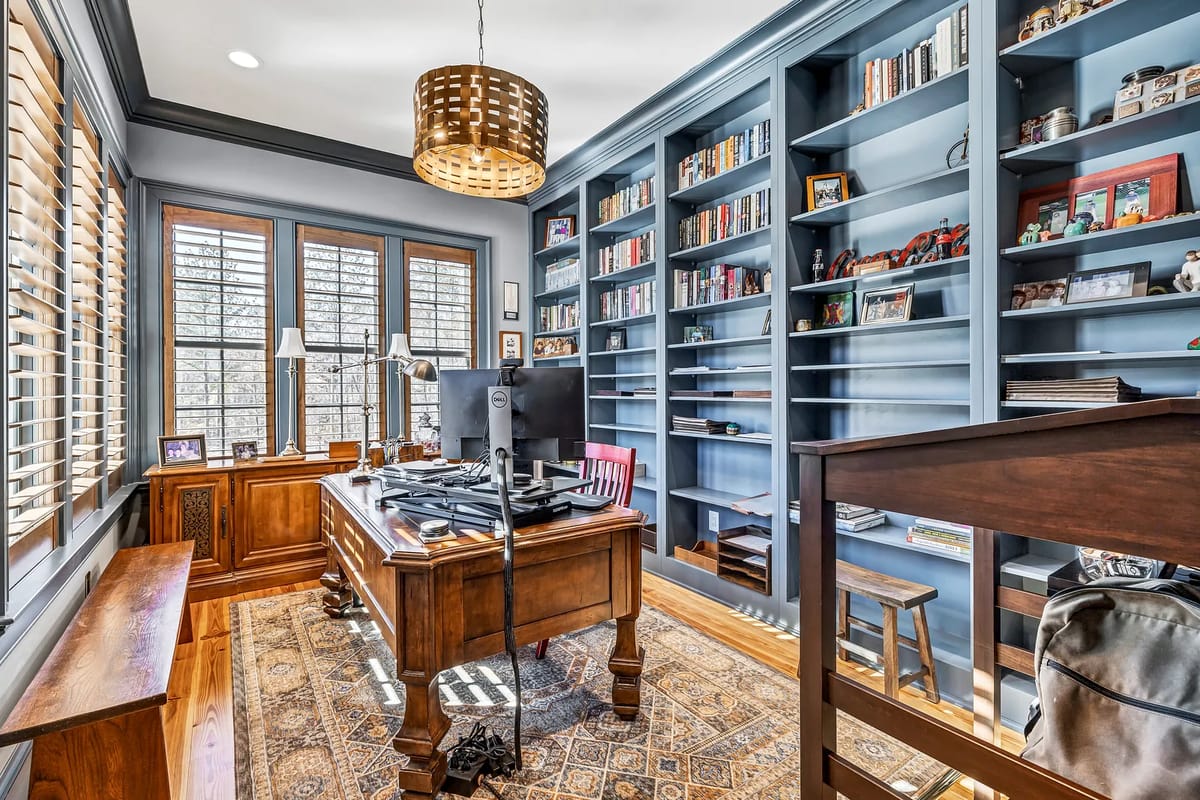Office with wooden desk, computer, bookshelves, two windows, and patterned area rug.