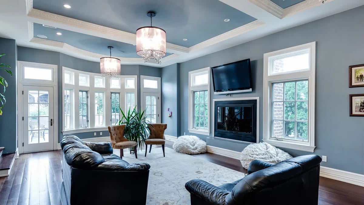 Living room with black leather sectional, patterned chairs, area rug, TV, windows, and detailed ceiling.