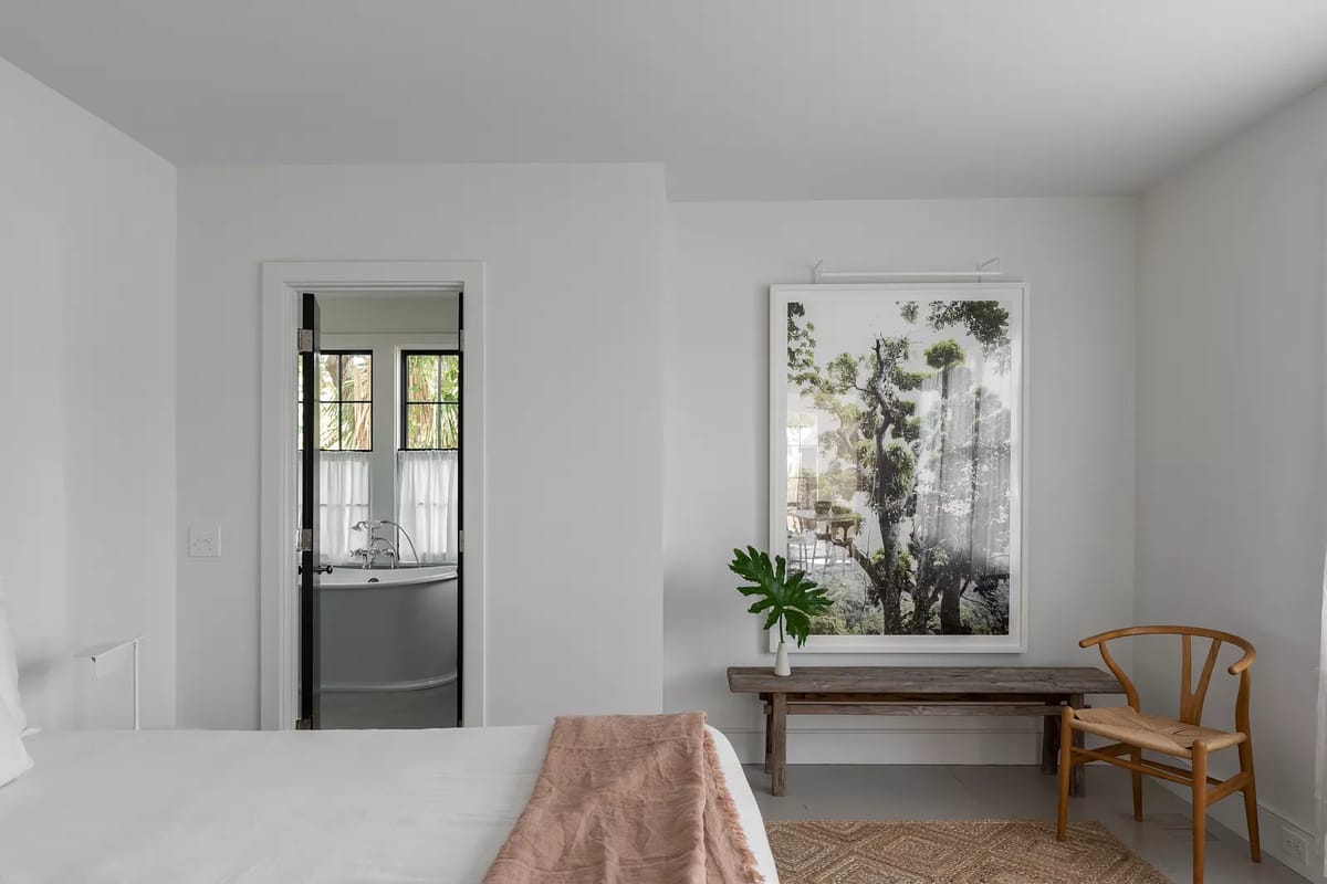 Bedroom with white bed, pink throw blanket, wooden bench, large nature artwork, and doorway to bathroom