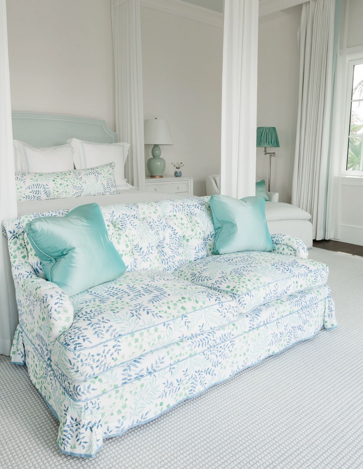 Bedroom with floral-patterned sofa, white bed linens, nightstands, and large windows with sheer curtains.