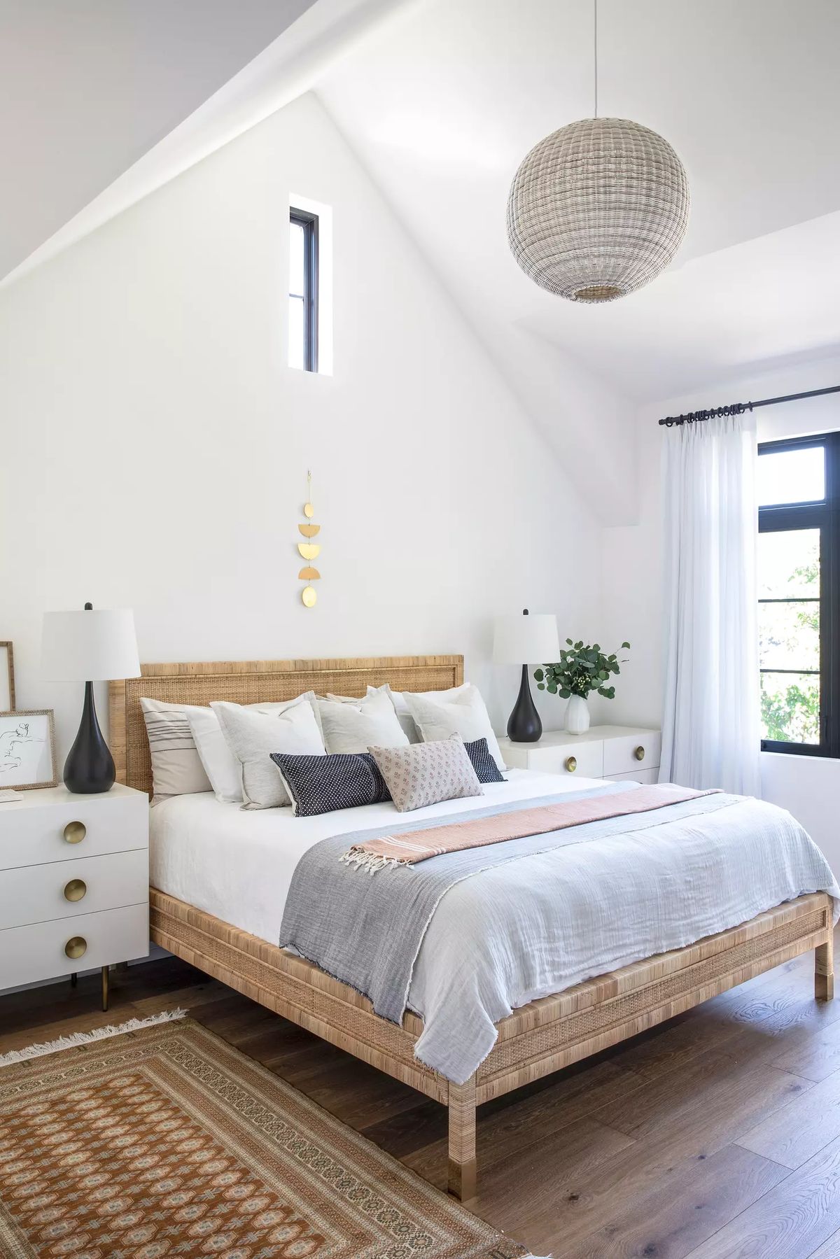 Bedroom with wooden bed, bedside tables, lamps, patterned rug, and large window with sheer curtains.