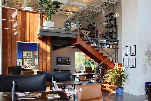Office space with workstations, wooden staircase, loft area, windows, plants, and artwork.