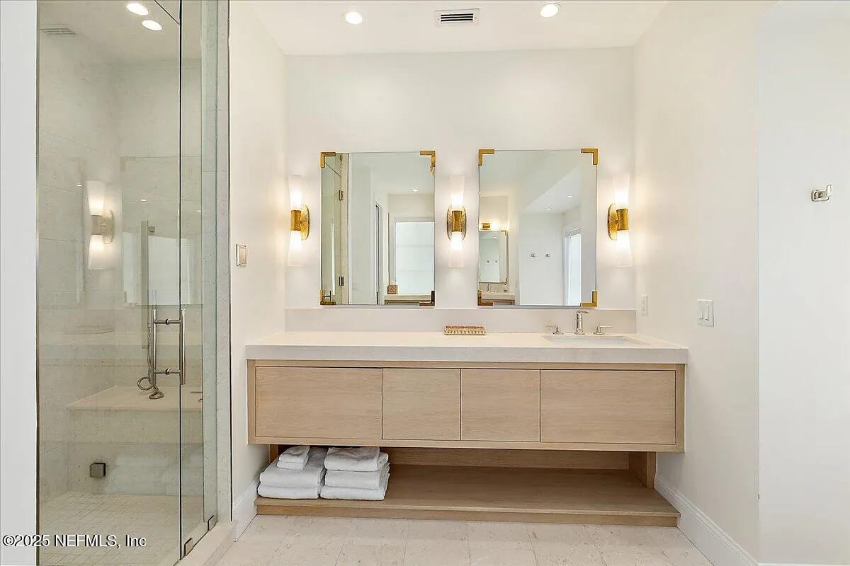 Bathroom with double sink vanity, mirrors, wall sconces, glass shower, and stacked towels.