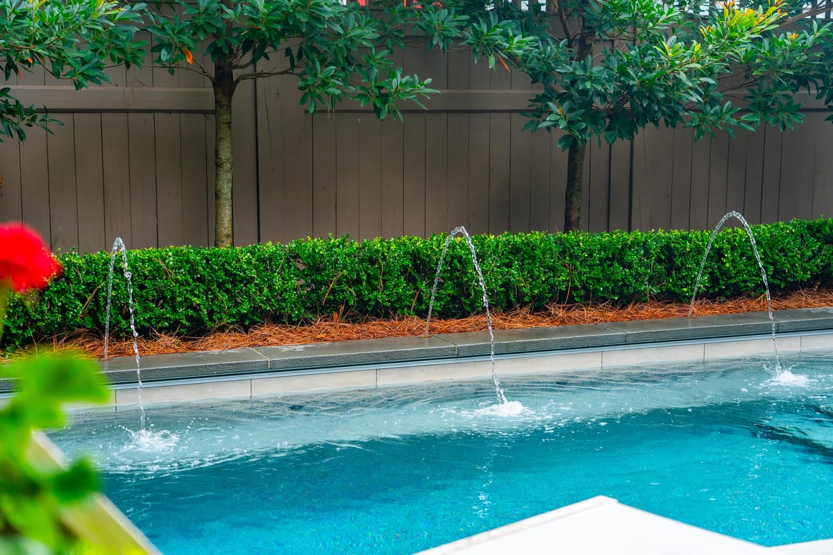 Garden area with swimming pool, water jets, hedges, and wooden fence.