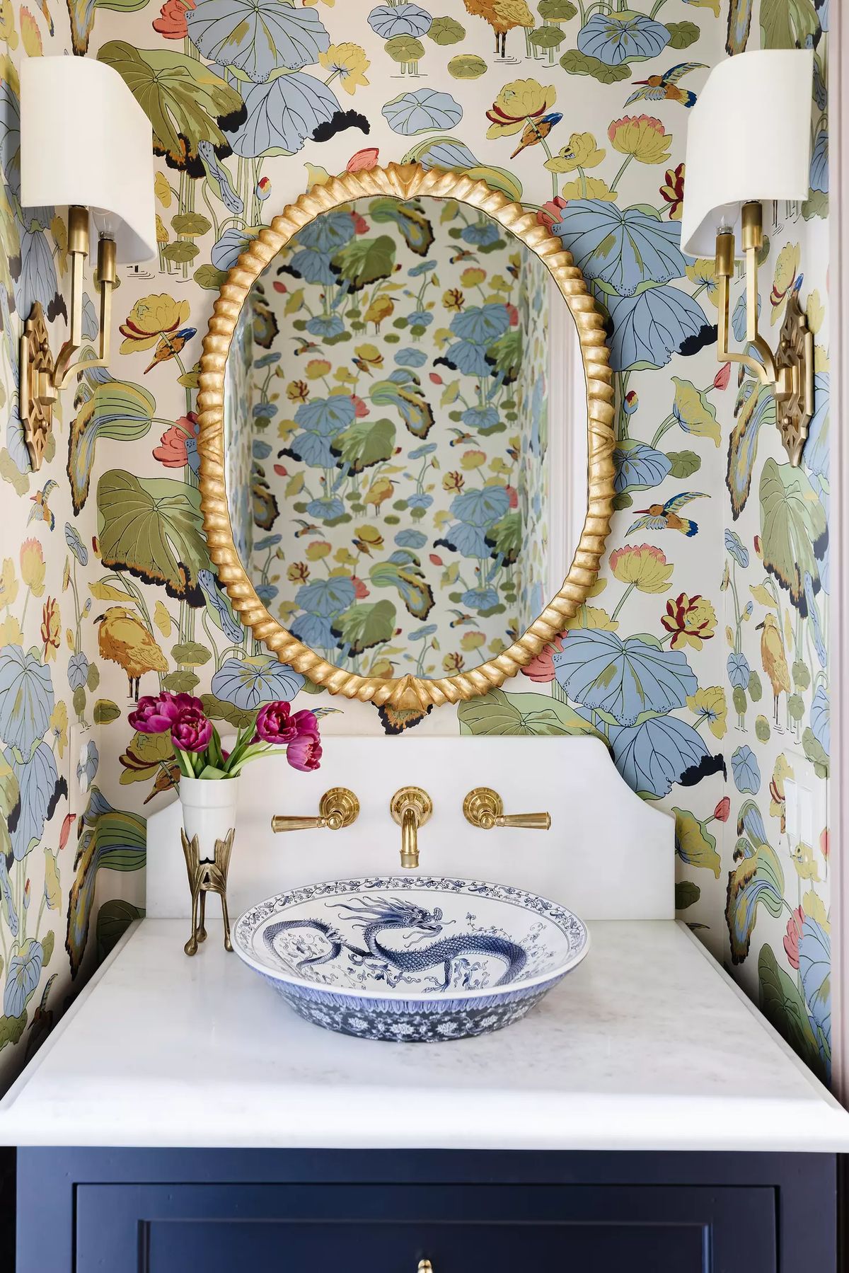 Powder room with floral wallpaper, round gold-framed mirror, marble countertop, blue and white sink, and wall sconces.