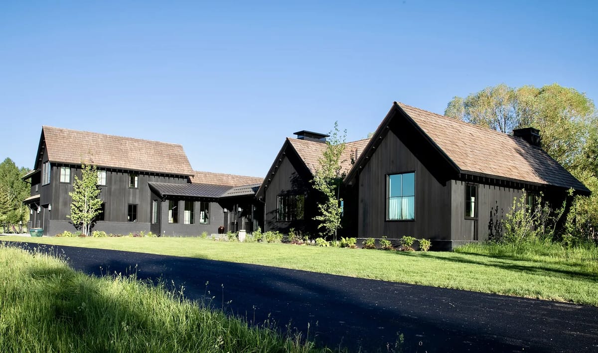 Front exterior of a black wood house with gable roofs, stone chimney, dark driveway, and landscaping.