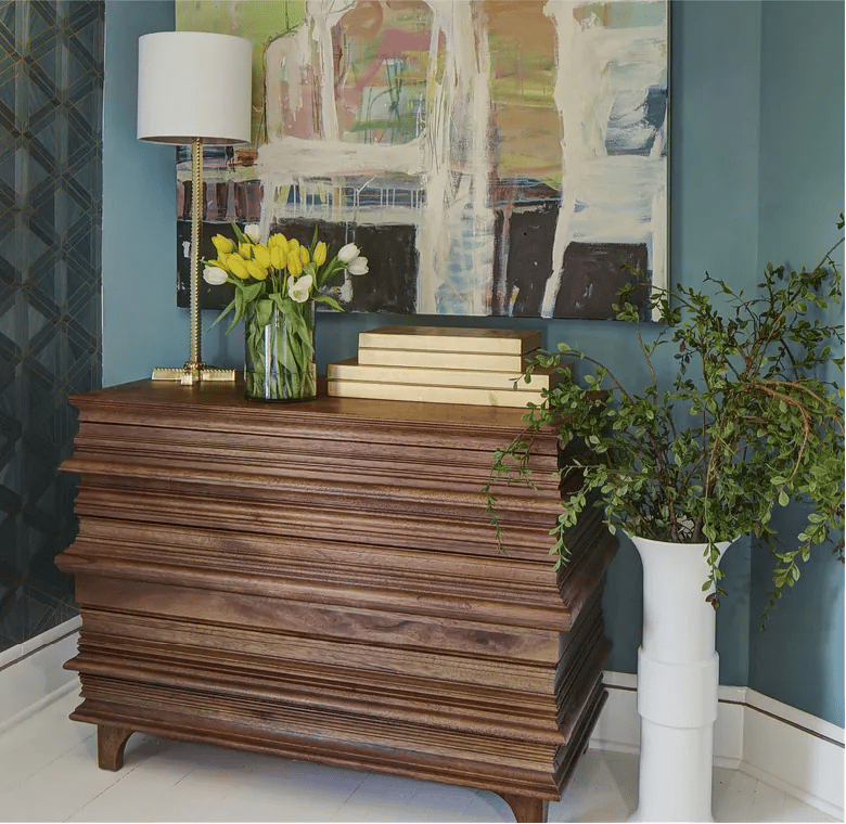 Styled corner with wooden sideboard, table lamp, tulips, books, and abstract artwork on blue wall.