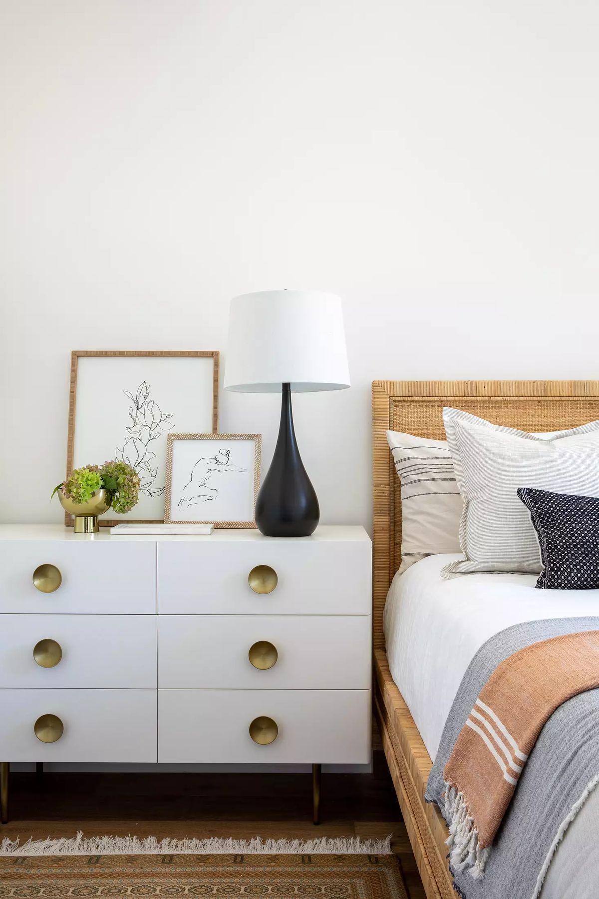 Bedroom with woven bed frame, white bedding, nightstand with black lamp and framed artwork, and cream area rug.