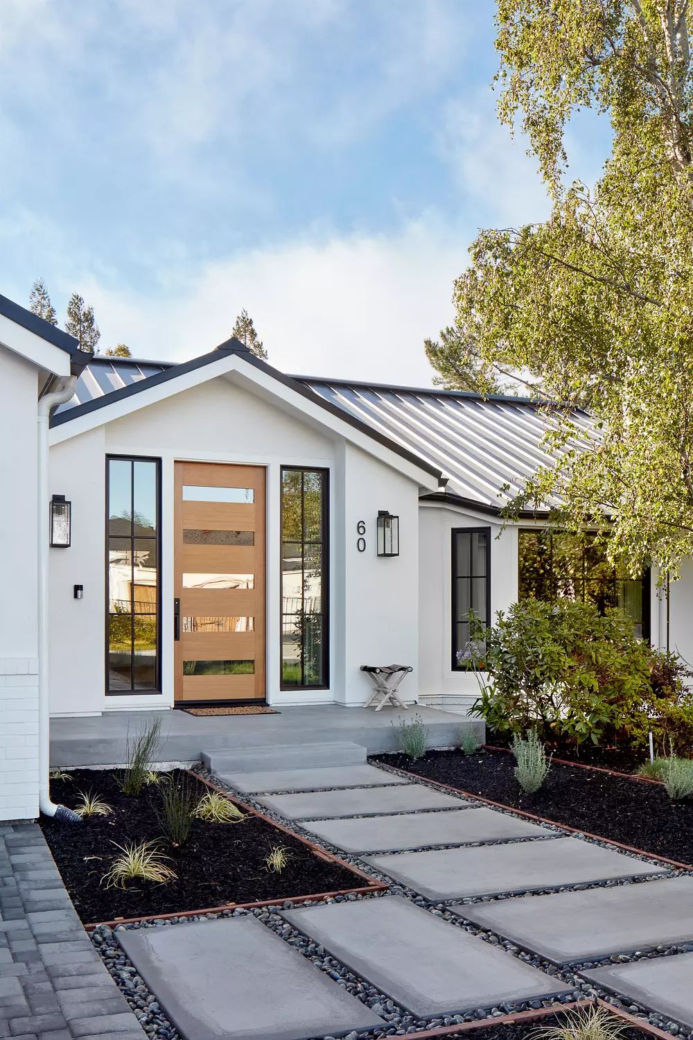Exterior front with wooden door, vertical windows, stone pathway, and small round table.