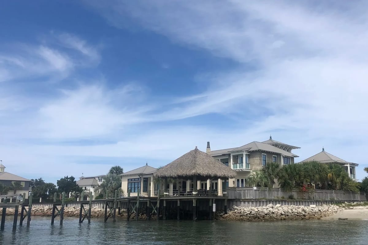 Coastal exterior view with thatched gazebo, wooden pilings, and landscaped greenery.