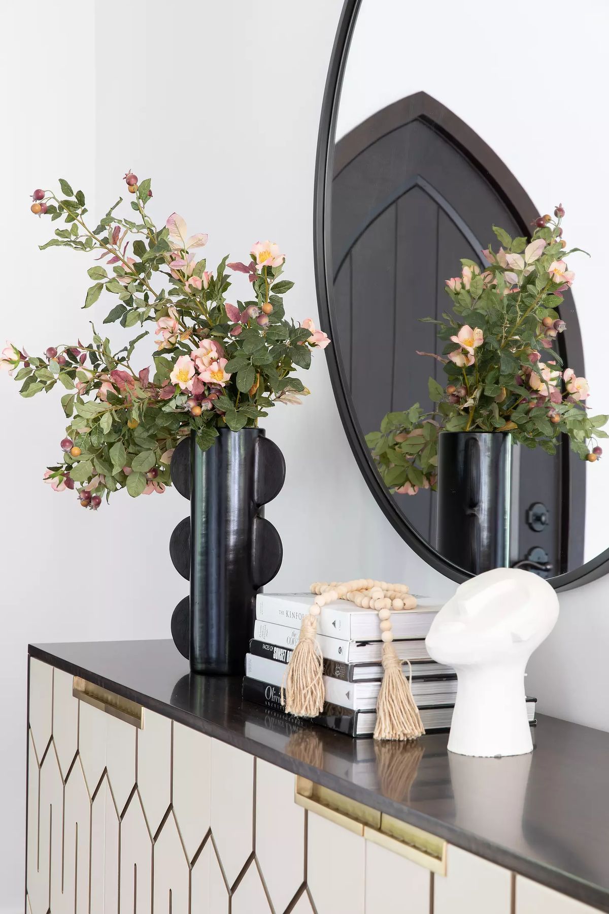 Foyer with black console table, round mirror, flowers, stacked books, and decorative object.