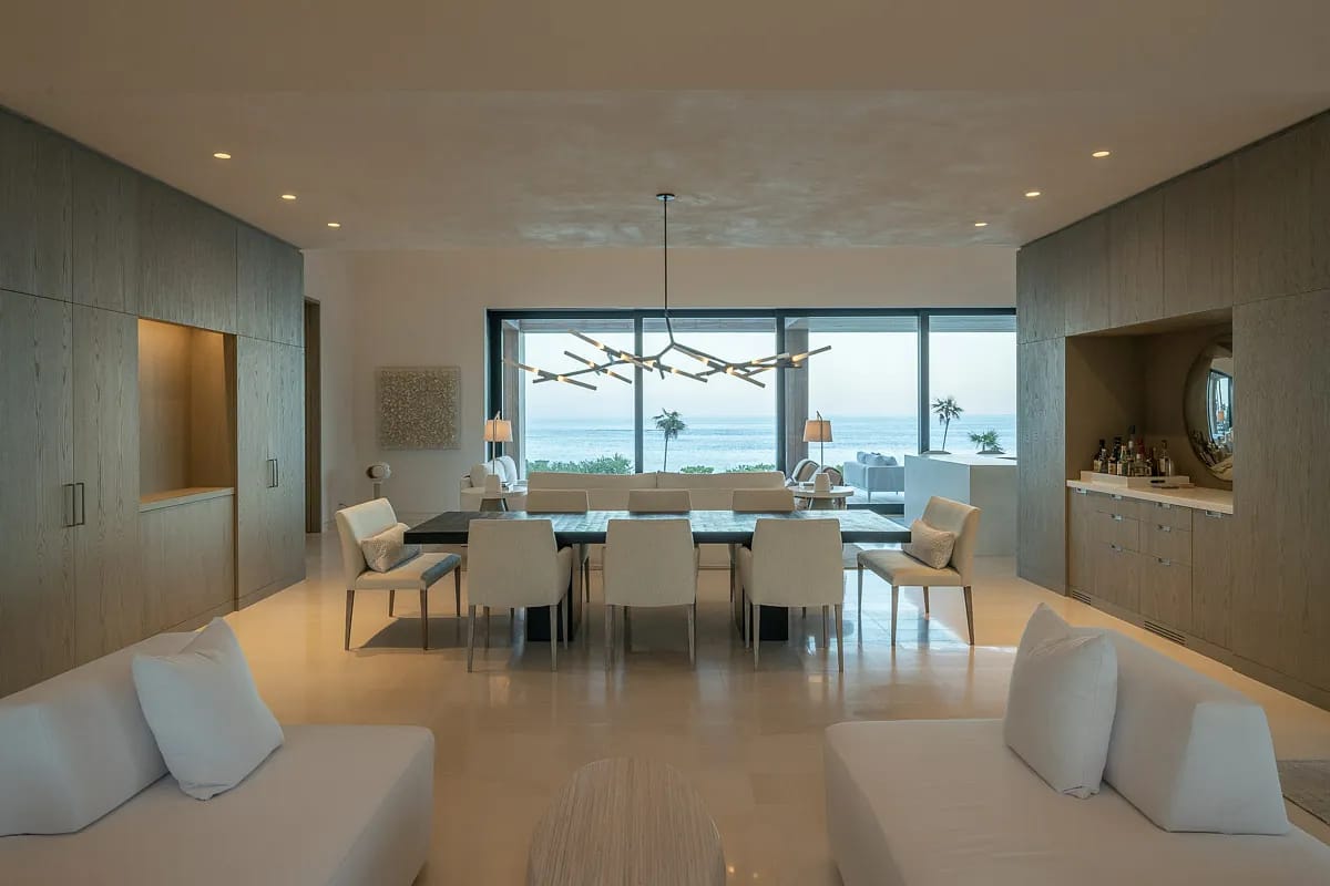 Dining room with long table, white chairs, modern chandelier, large windows, and bar area with cabinetry.