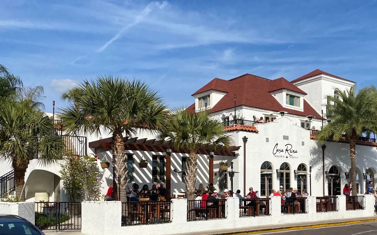 Restaurant exterior with white stucco, red tiled roof, palm trees, and outdoor seating under a pergola.