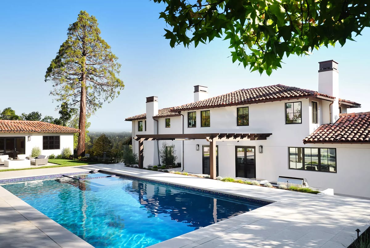 Exterior rear view of a house with a swimming pool, stone paving, white walls, red tile roof, and landscaping.