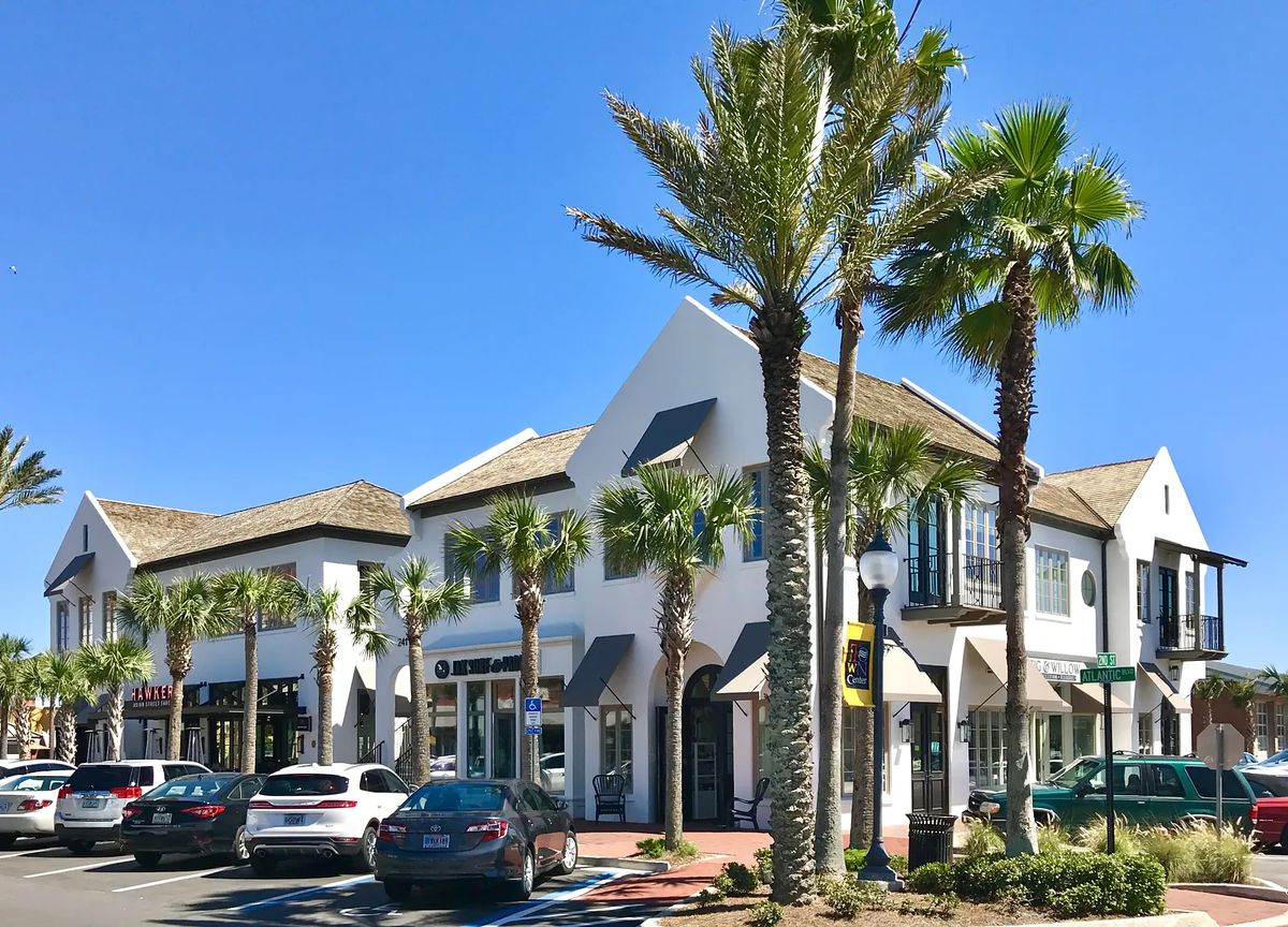 Exterior view of a building with shops, white walls, brown roof, palm trees, and parked cars.