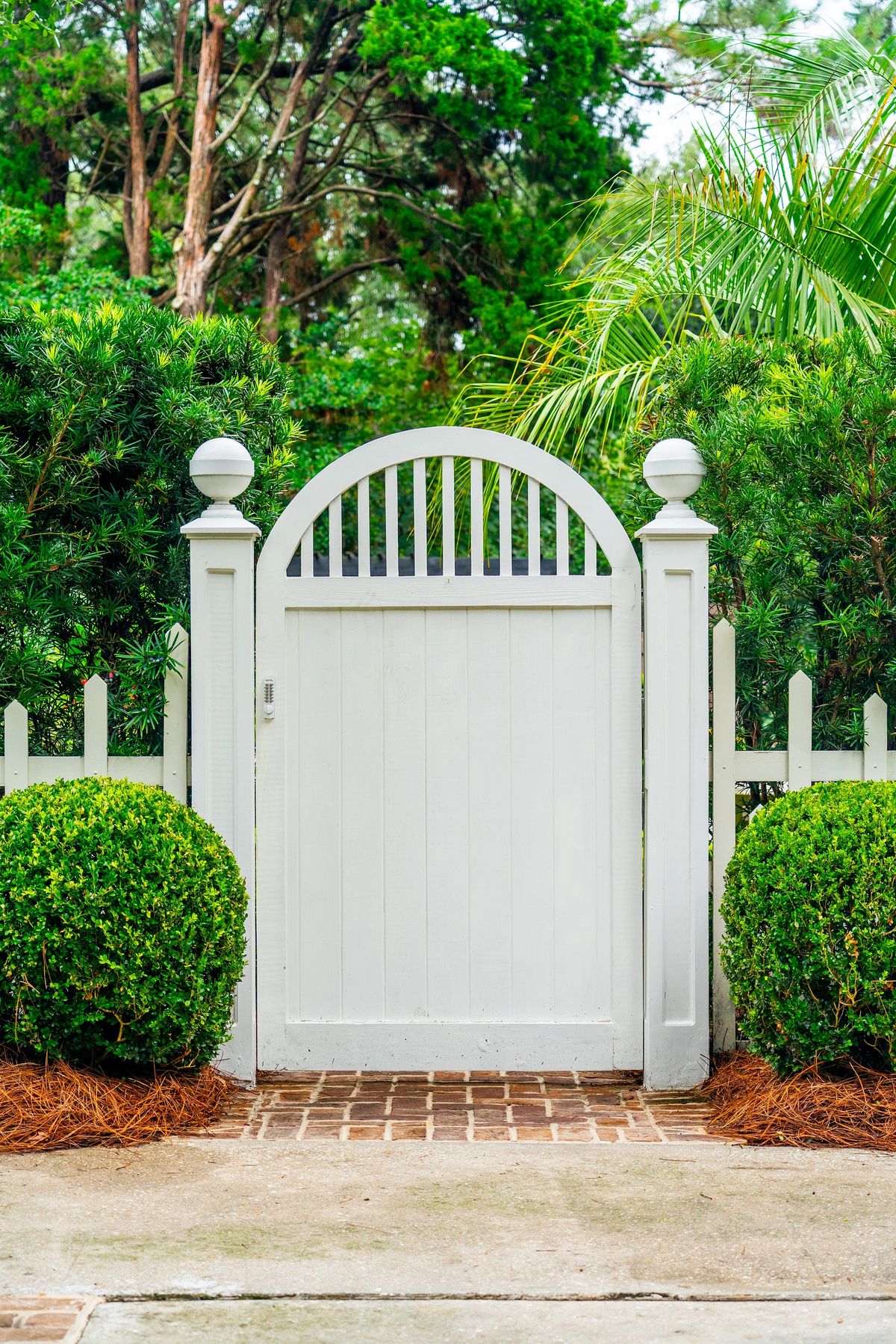 Exterior garden gate with white wooden structure, brick path, and manicured bushes.