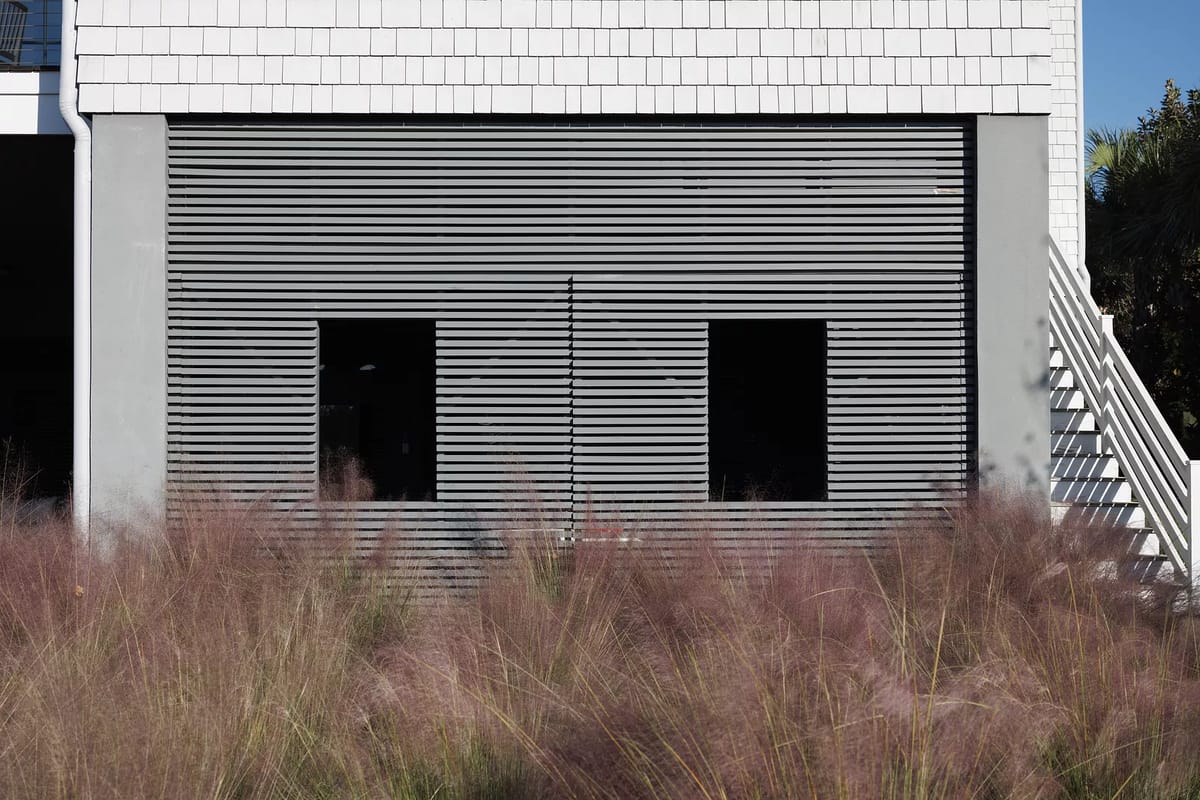 Exterior view of building with wood slats over windows, pink grass, and visible staircase.