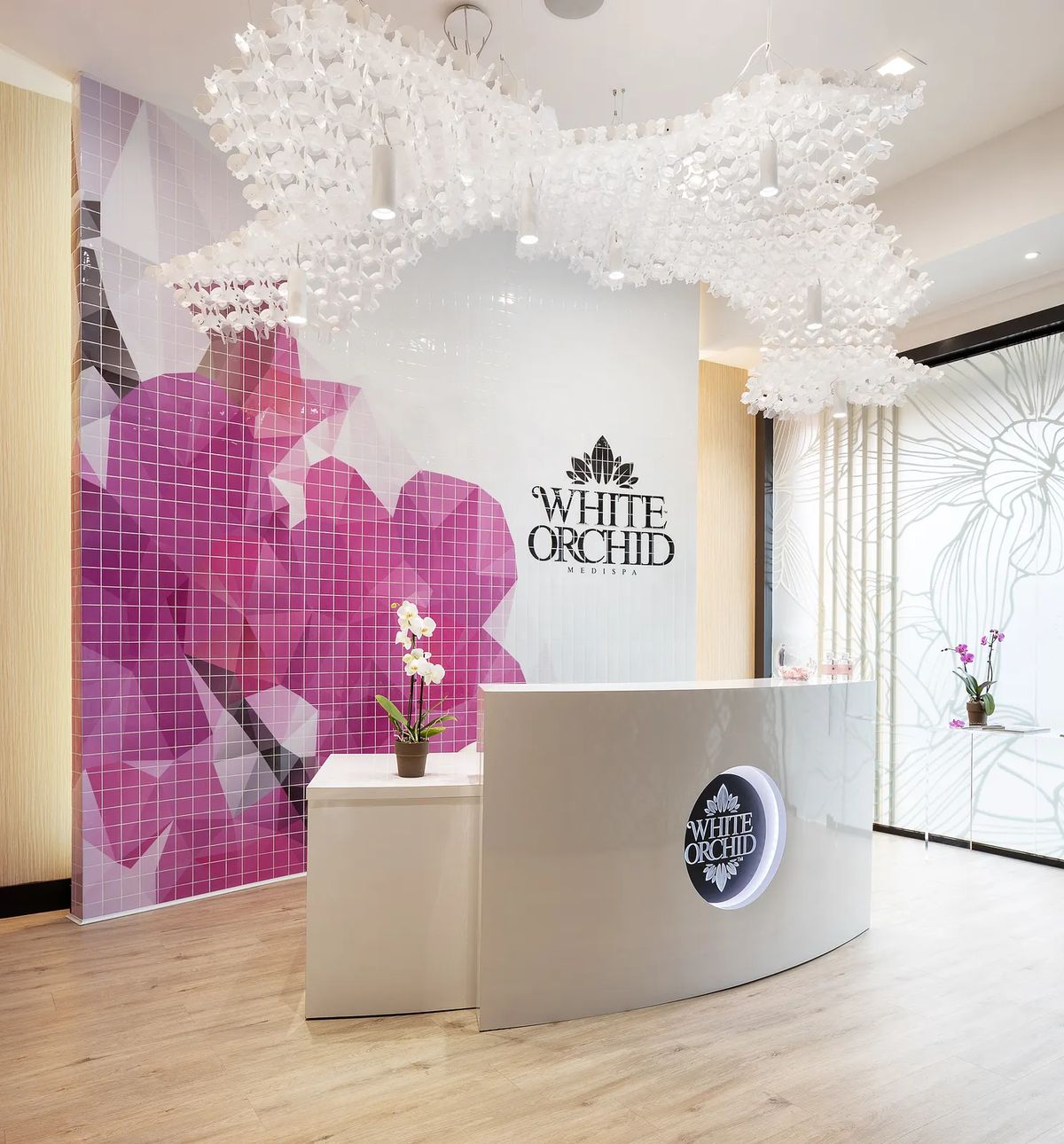Reception area with white desk, pink and purple mural, orchids, light wood floor, and unique chandelier.