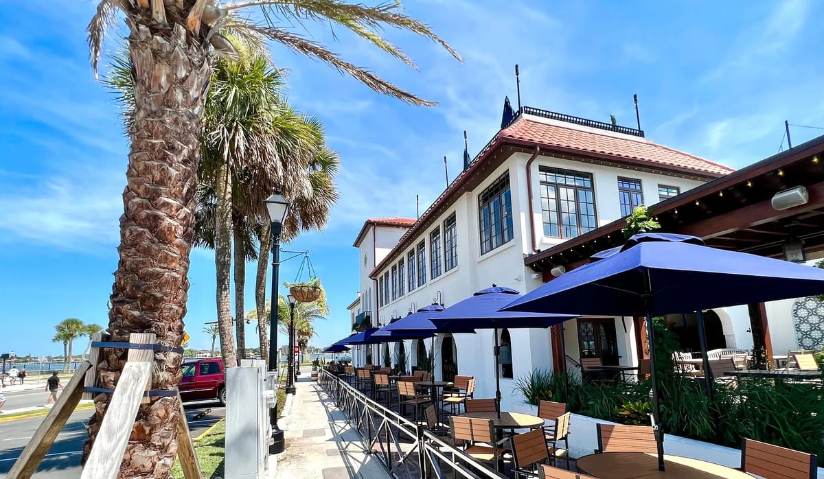 Exterior view of a building with palm trees, outdoor seating, blue umbrellas, and a walkway.