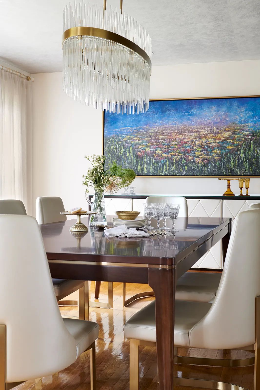 Dining room with dark wood table, white chairs, wall painting, flowers, and chandelier above wooden flooring