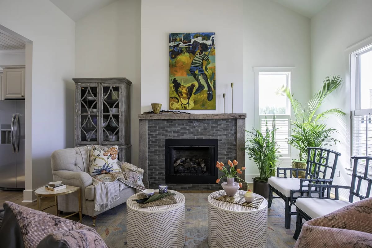 Living room with gray stone fireplace, painting, sofa, chairs, coffee tables, and tall plant with natural light.