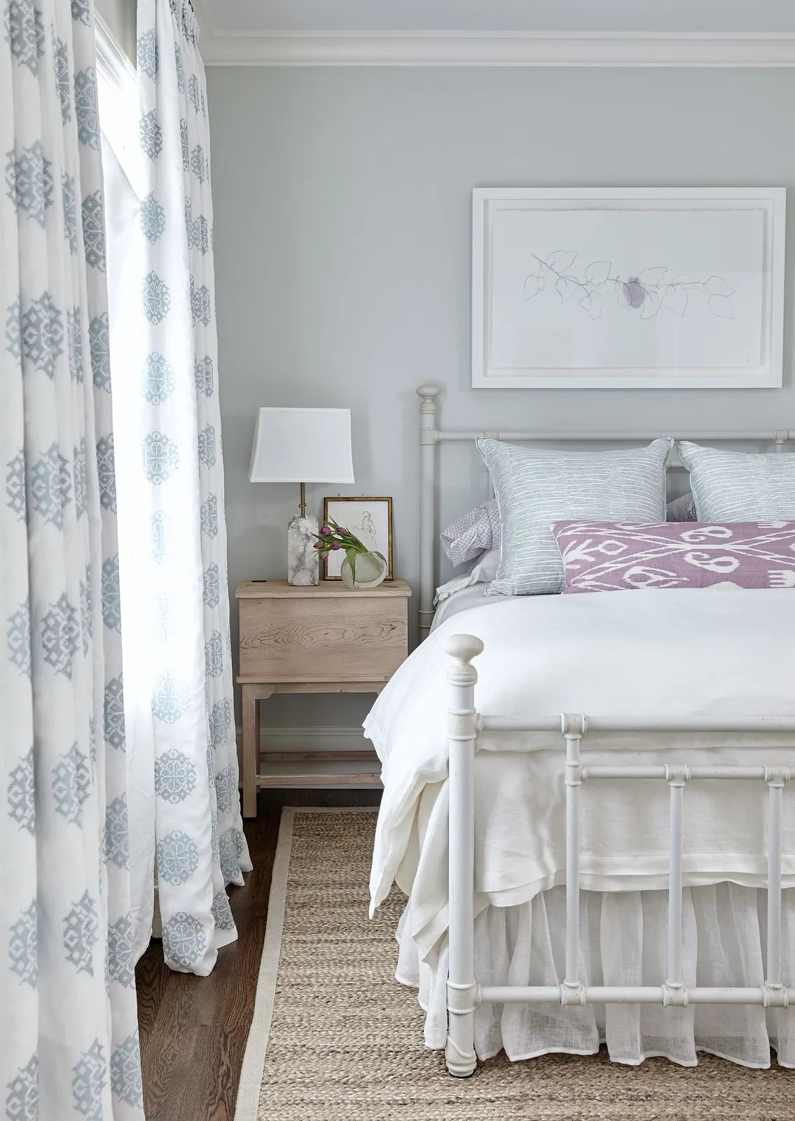 Bedroom with metal bed frame, white linens, nightstand with lamp, light blue curtains, and jute rug.