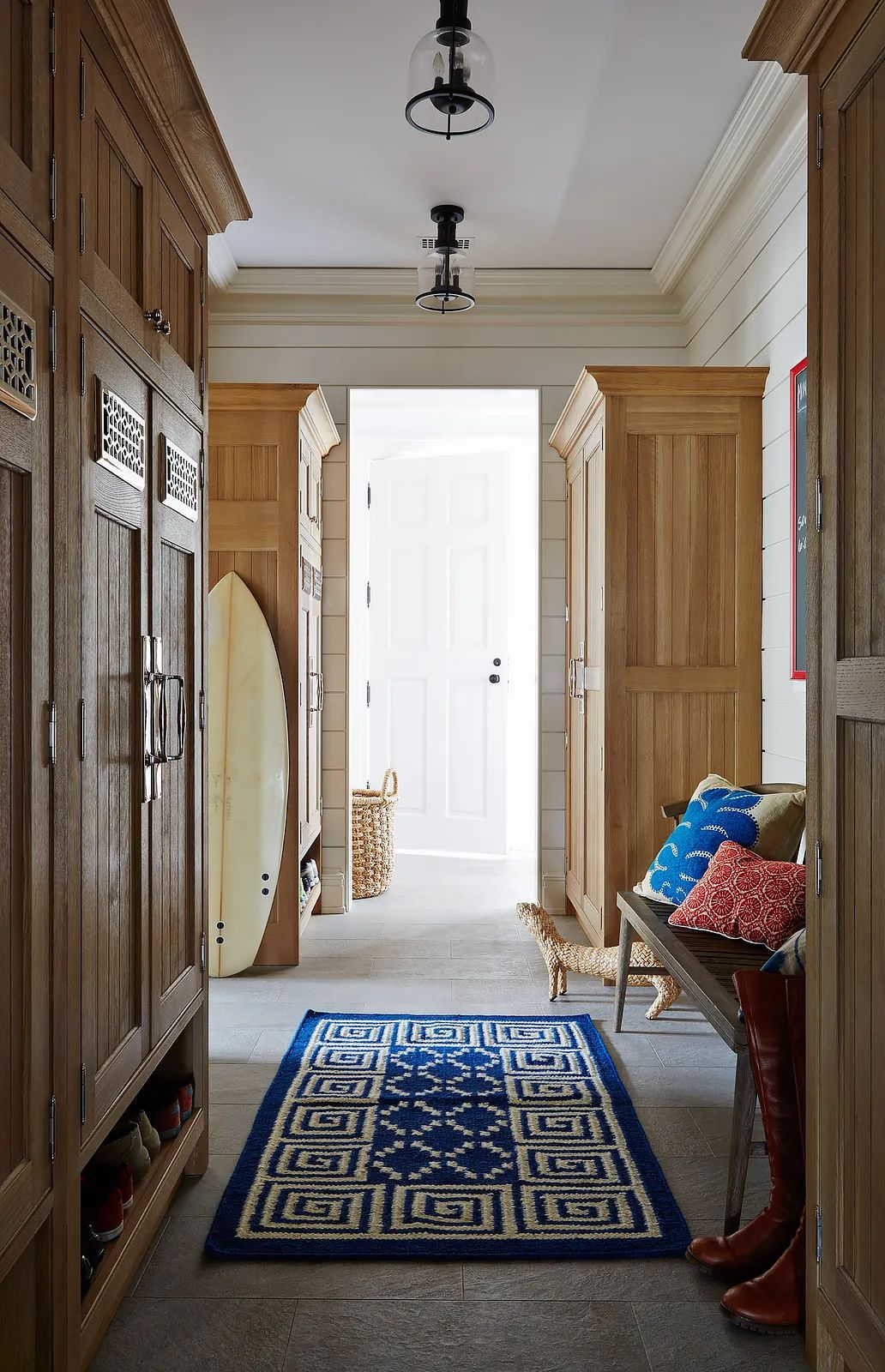 Mudroom with wooden lockers, blue rug, bench, surfboard, and light-colored walls with natural light.