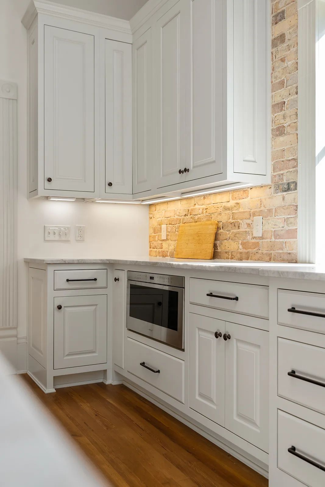 Kitchen with white cabinets, built-in microwave, wooden cutting board, brick backsplash, and hardwood floor.