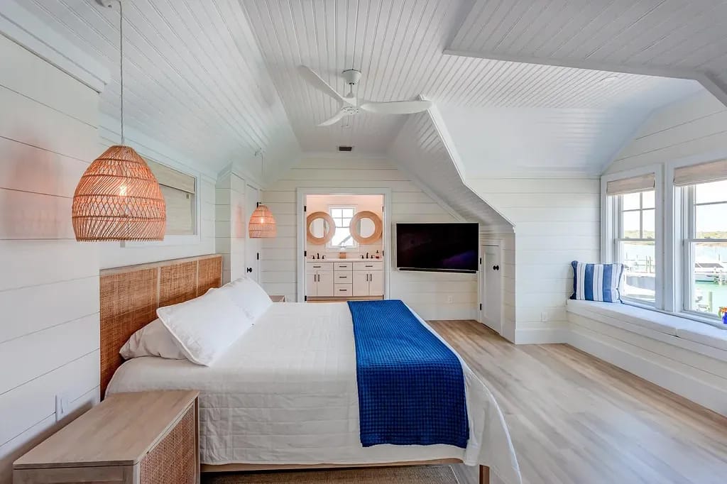 Bedroom with white bed, blue blanket, woven lights, TV, and large windows.