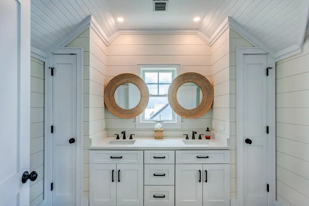 Bathroom with dual sinks, round mirrors, white paneled walls, and two doors.
