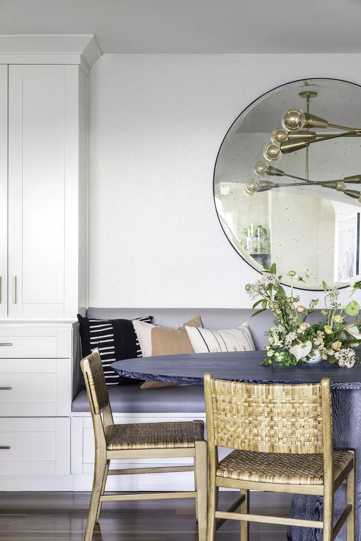 Dining room with round table, woven chairs, floral arrangements, and large circular mirror.