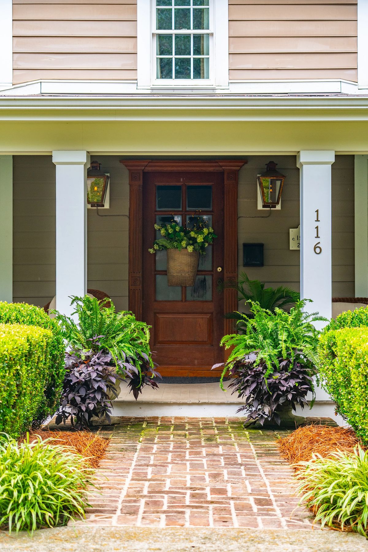 Exterior front with wooden door, lanterns, ferns, shrubs, and brick pathway.