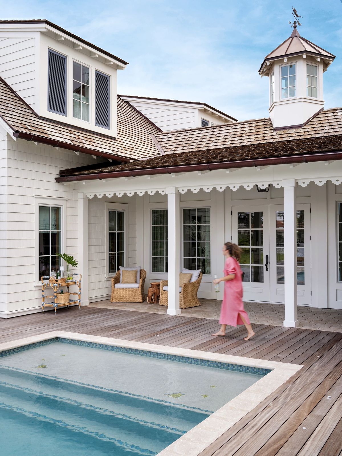 Patio with pool, wooden decking, wicker chairs, side table, and house with windows and cupola