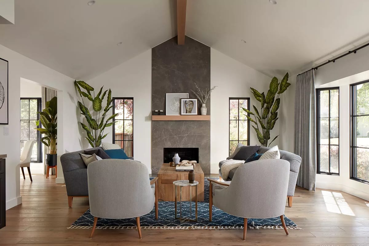 Living room with gray armchairs, gray sofa, wood coffee table, stone fireplace, and large windows.