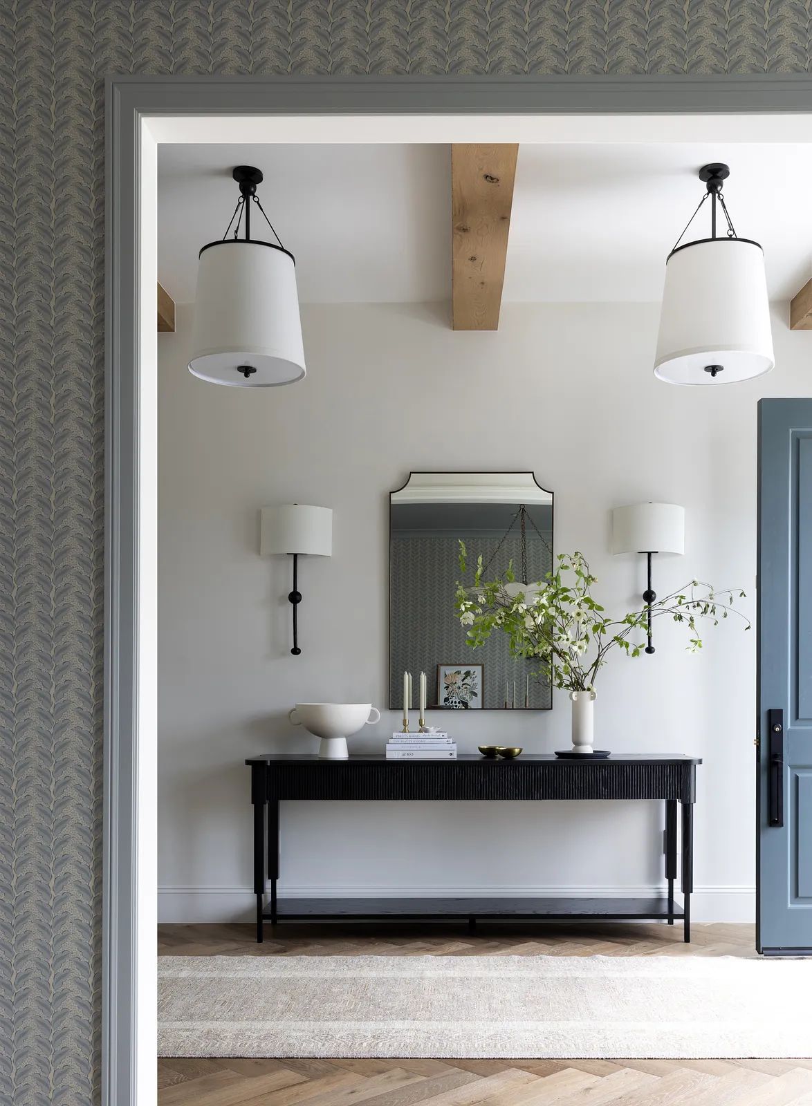 Foyer with black console table, wall mirror, sconces, neutral rug, and decorative bowl with branches.