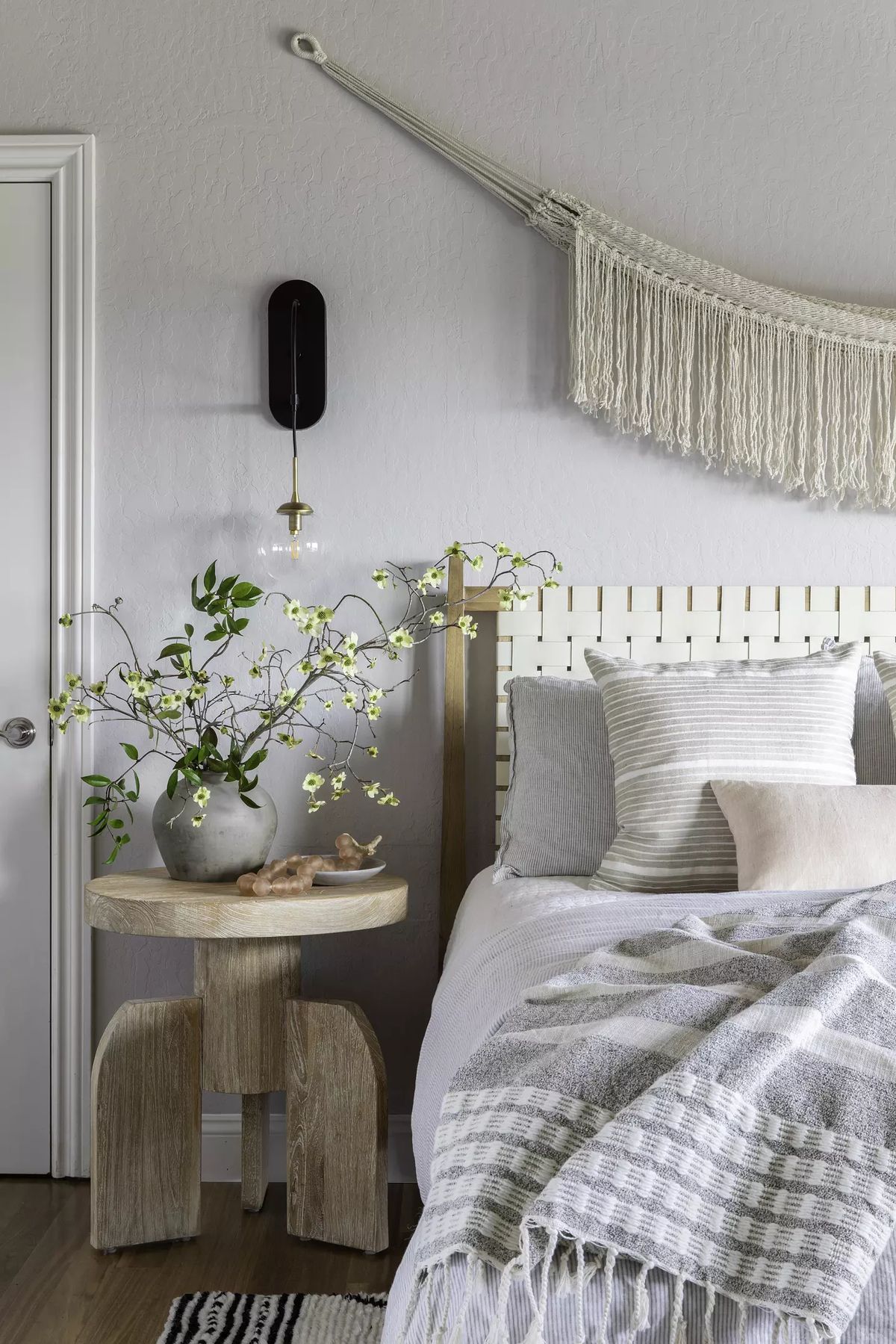 Bedroom with gray bedding, wood side table, vase of flowers, and wall hanging.