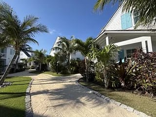 Exterior view with sandy pathway, palm trees, colorful shrubs, and houses with white siding and blue shutters.