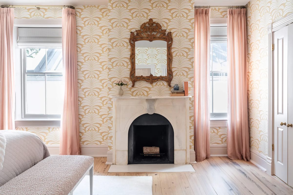 Bedroom with decorative fireplace, mirror, pink curtains, patterned wallpaper, and a bench.