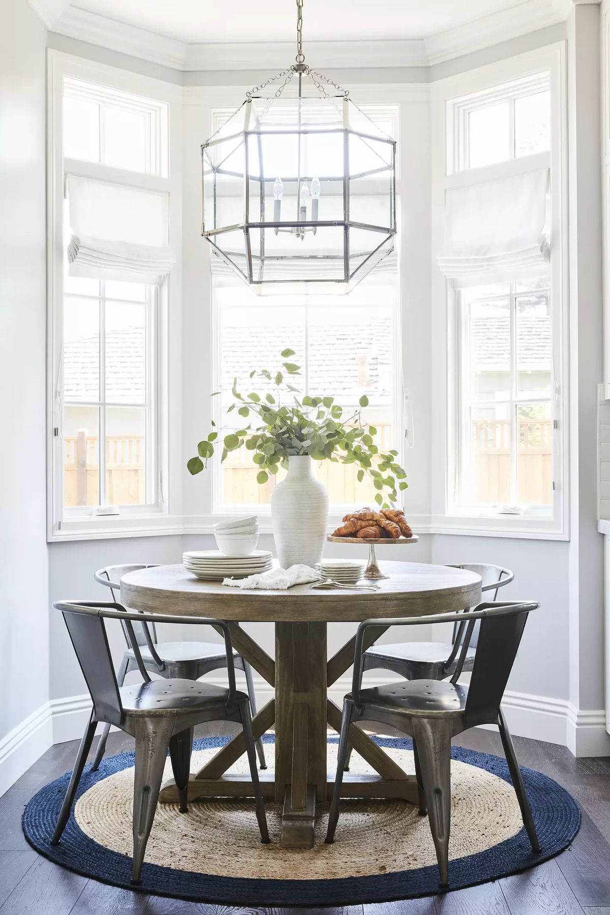 Dining room with round wooden table, black chairs, greenery vase, pendant light, and large windows
