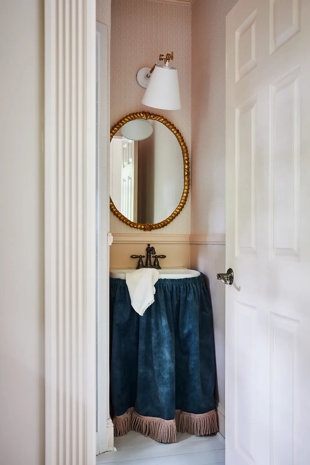 Powder room with round gold-framed mirror, dark blue sink skirt, light patterned wallpaper, and wall sconce.