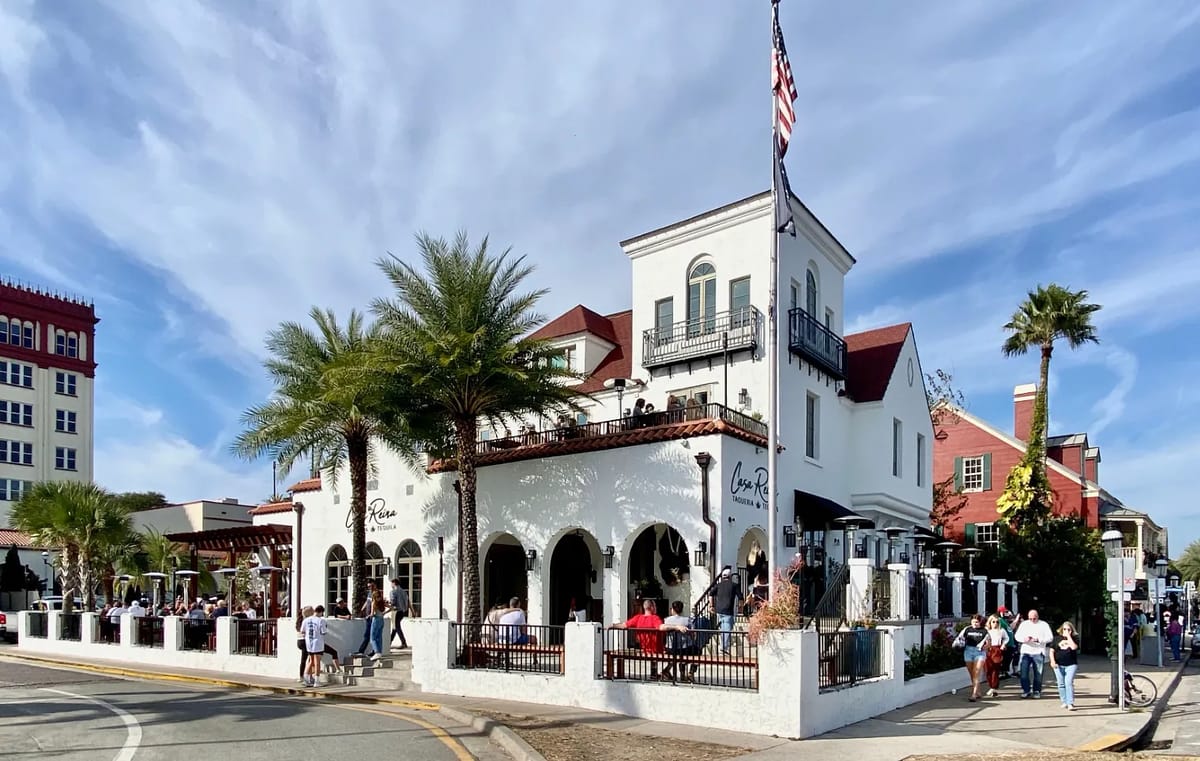 Restaurant exterior with white stucco walls, arches, red roof, palm trees, outdoor seating, and balcony.