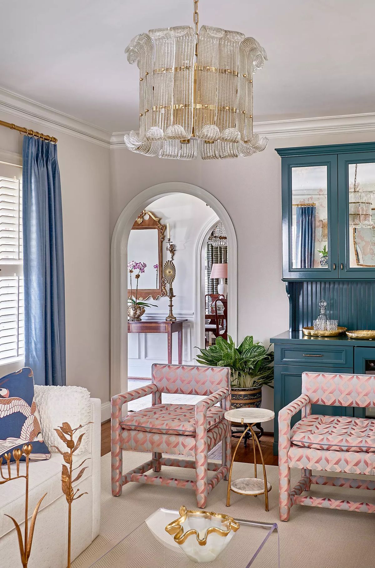 Living room with white sofa, pink armchairs, gold side table, blue drapes, and decorative mirror.