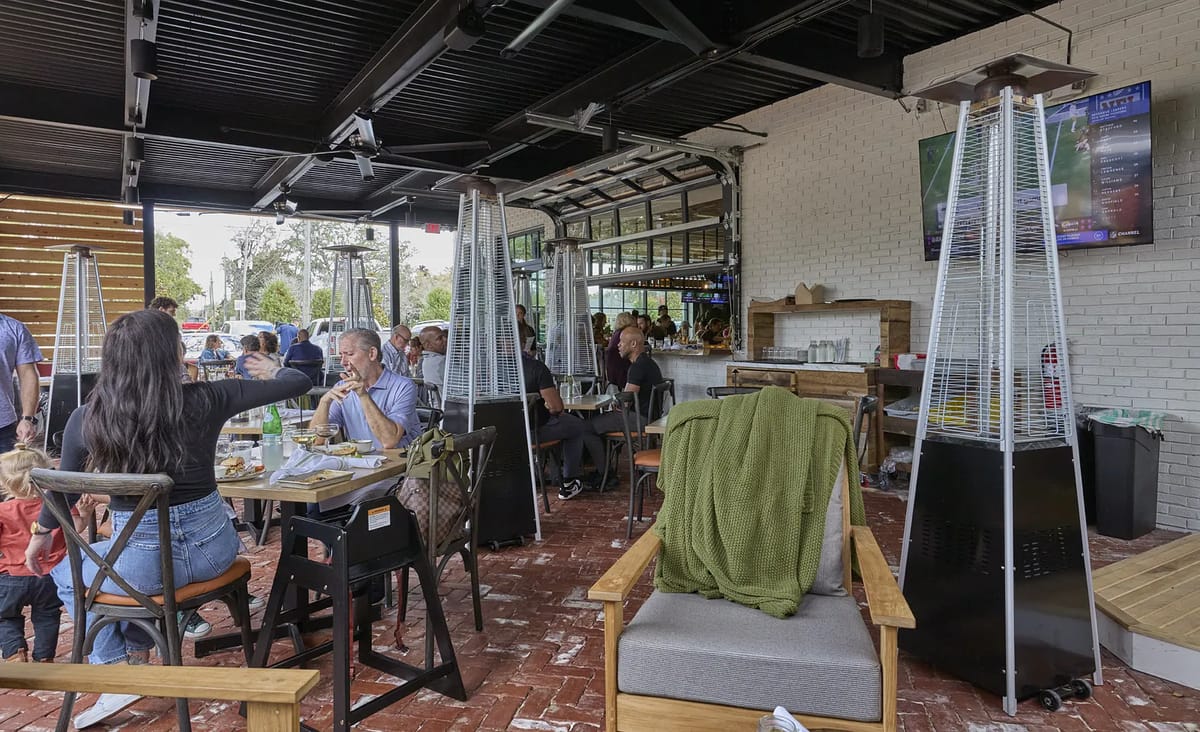 Outdoor patio with wooden chairs, glass tables, heat lamps, and a visible bar area.