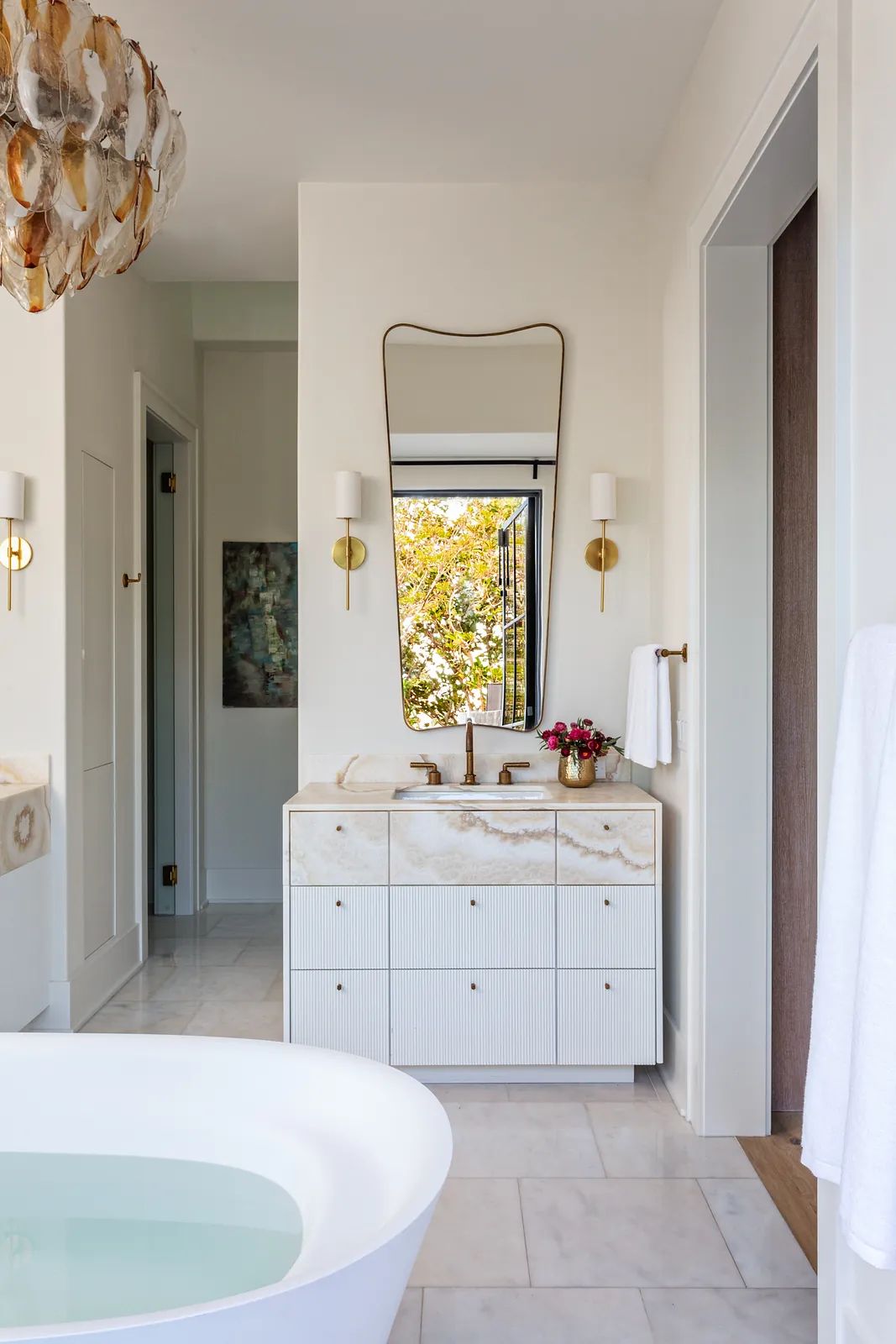 Bathroom with freestanding white bathtub, marble vanity, mirror, two sconces, and large white tiles.